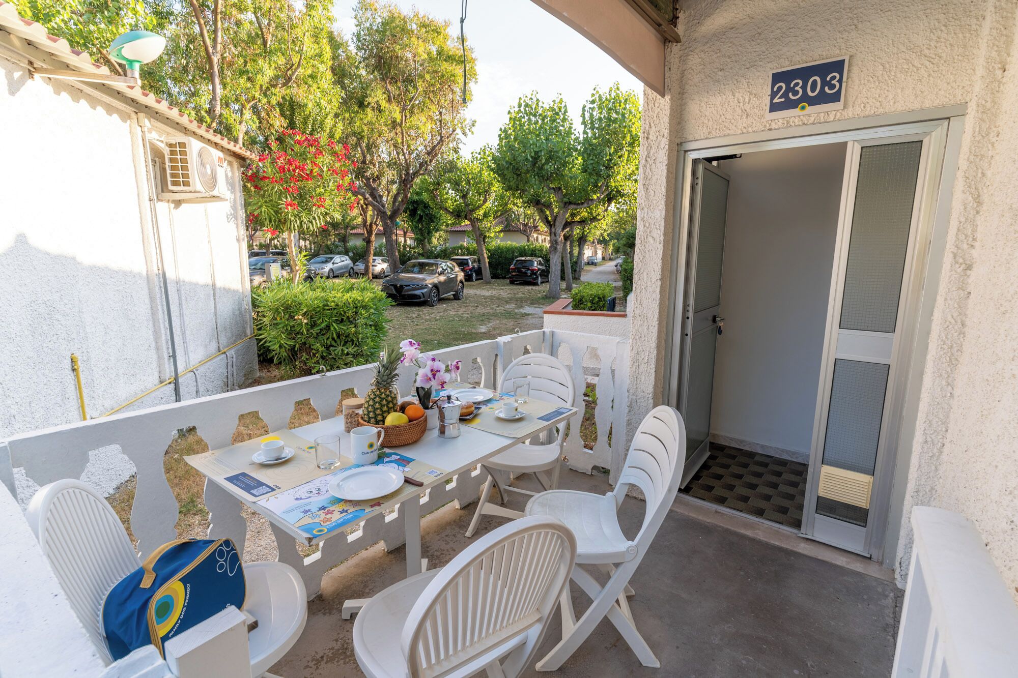 A balcony with a table set for breakfast, white chairs, a small table with a basket of fruit, and a door with the number 2303.