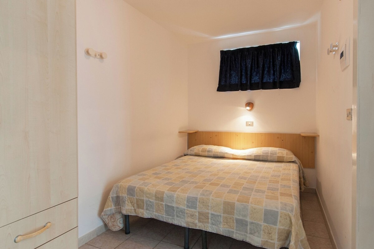 A small, single-bed room with a wooden headboard, a bedside lamp, and a window with a dark curtain.