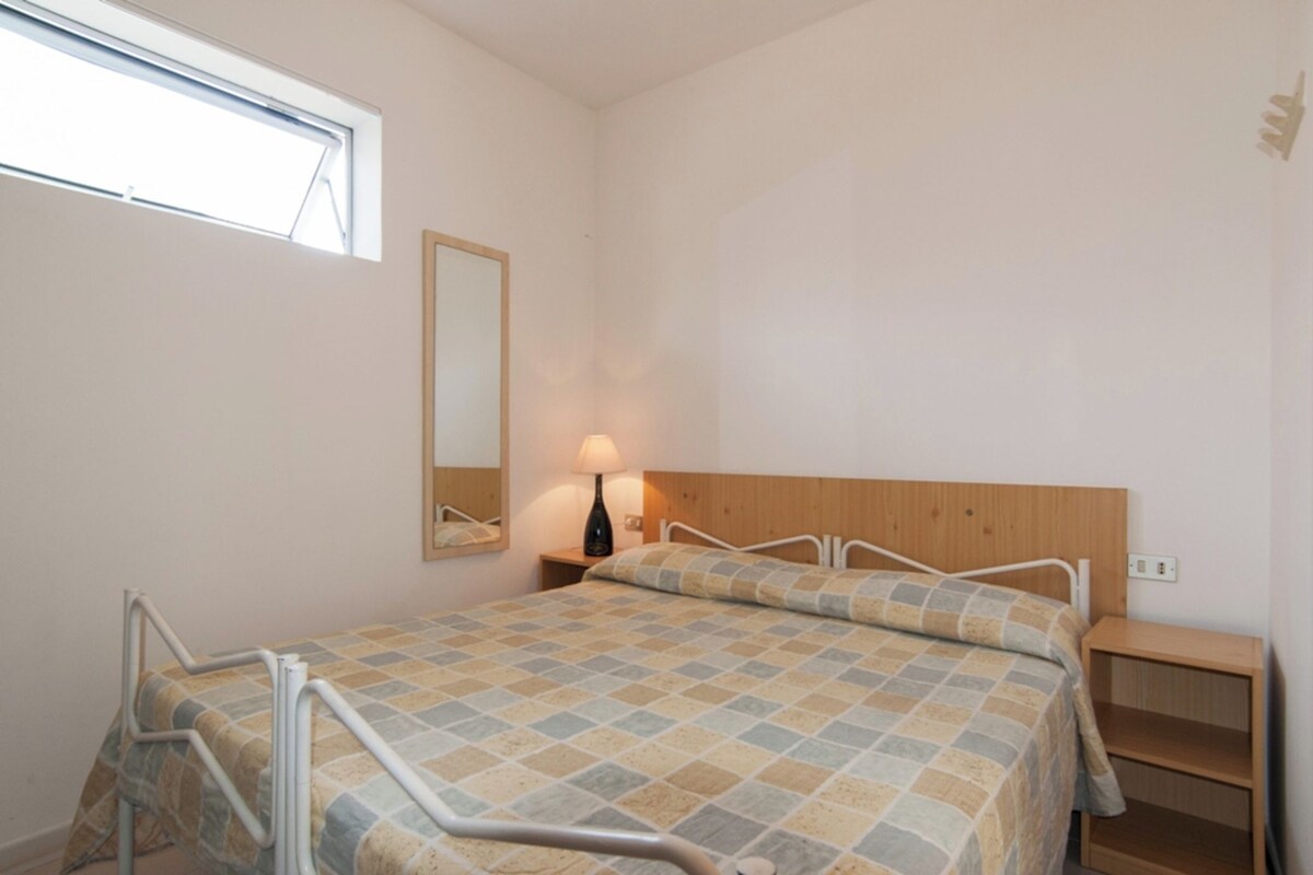 A bedroom with a bed, a nightstand, a lamp, a wooden headboard, a window, and a mirror.