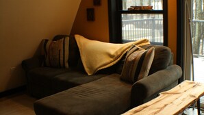 Ein modernes Wohnzimmer mit einem Ecksofa, einem Holztisch und einem großen Fenster mit Blick auf Bäume.