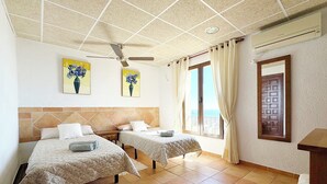 Quarto de hotel com duas camas, um ventilador de teto, uma janela com cortinas e uma porta com padrão geométrico.