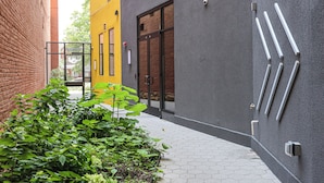 A narrow alley with a yellow building, a grey building, and a brick wall. There are plants in the foreground and a pathway with hexagonal tiles. - Studio Apartment in the Heart of Philadelphia. (Philadelphia)