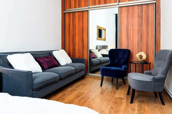 A modern living room with a grey sofa, a blue armchair, and a grey armchair. There is a wooden wardrobe with a mirror and a small round table with a decorative item.