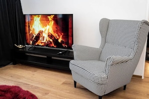 A living room with a black houndstooth patterned armchair, a flat-screen TV displaying a fireplace, and a wooden floor.