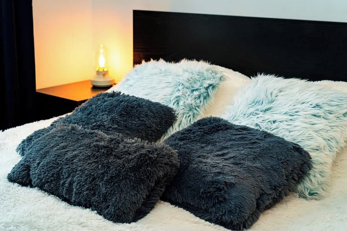 Een bed met zachte kussens in blauw- en grijstinten, een witte, zachte deken en een nachtkastje met een lamp.