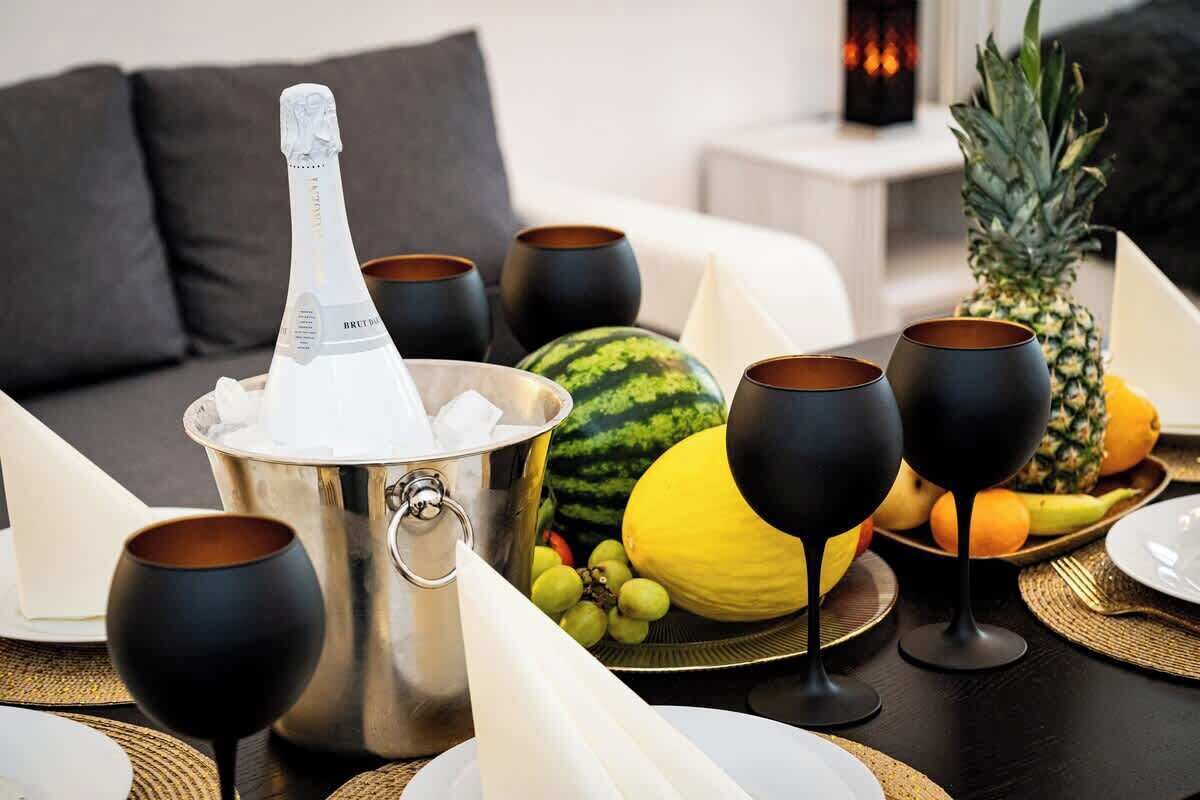 Una mesa con servicio de cena, una botella de champán, copas de vino negras y un plato de frutas.