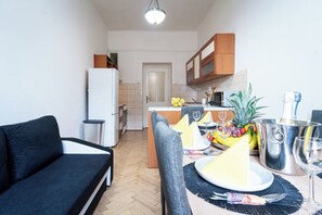 Un espacio habitable compacto con sofá, mesa de comedor para dos personas, cocina con armarios y refrigerador, y una puerta que conduce a otra habitación.
