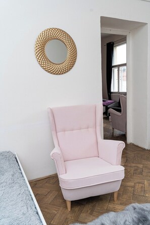 A room with a light pink armchair, a round mirror with a textured frame, and a view of a living area with a grey sofa and a window.