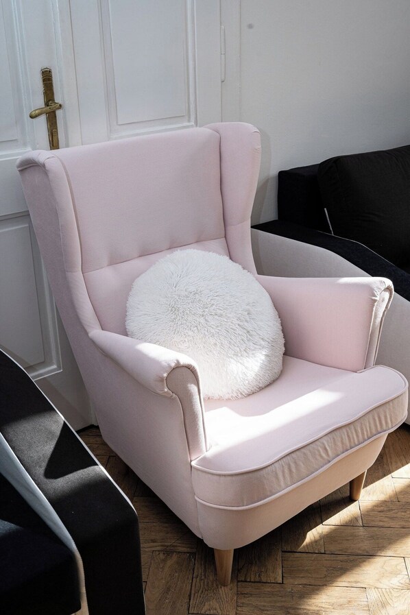 Un sillón rosa claro con un almohadón blanco mullido, situado en una habitación con suelo de madera y una puerta blanca al fondo.