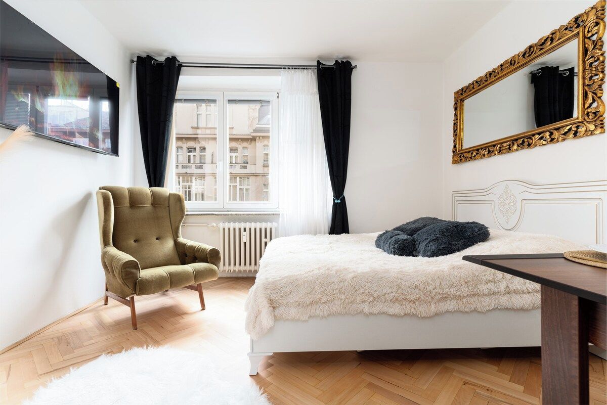 Une chambre à coucher avec un lit, une chaise, un grand miroir et une fenêtre avec des rideaux.