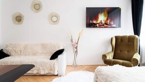 A living room with a white sofa, a brown armchair, a white furry rug, a wooden coffee table, and a painting of a fire on the wall. - Grand Luxury Suite Su (Prague)