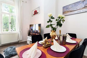 A dining area with a wooden table set for two, a vase with flowers, a champagne bottle, and a painting on the wall.