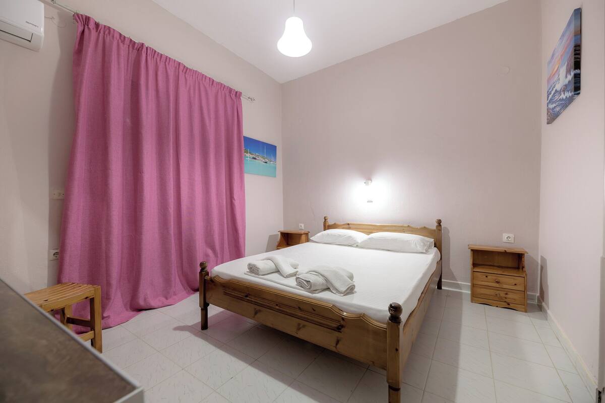 A bedroom with a wooden bed, a pink curtain, a small wooden nightstand, and a wall-mounted light.