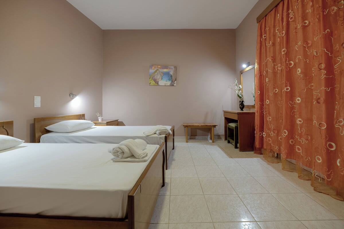 A hotel room with two beds, a small table, a chair, a desk, a mirror, and a painting on the wall.