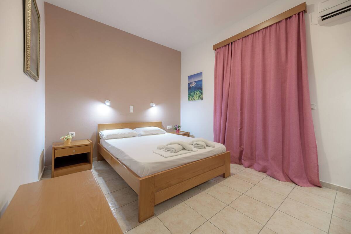 A bedroom with a wooden bed, bedside tables, a nightstand, a wall-mounted air conditioner, and a window with pink curtains.
