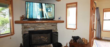A living room with a fireplace, a television on a wooden shelf, a stone wall, and a wooden floor.