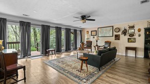 A spacious living room with hardwood flooring, a large window with curtains, a ceiling fan, and a variety of wall decorations including framed pictures and trophies.