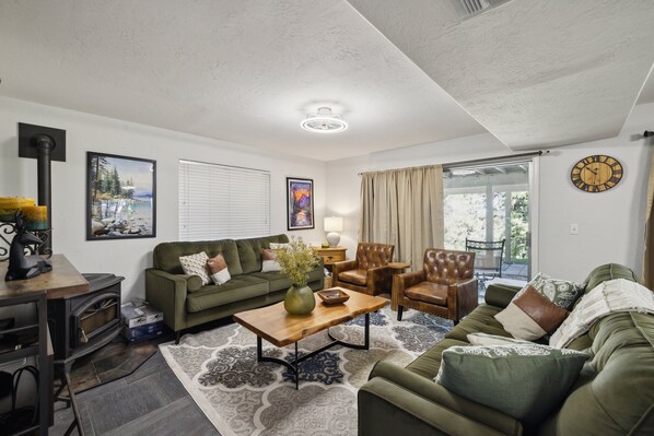 A living room with a fireplace, green sofas, a coffee table, and a large window with curtains. - Whispering Pines Retreat 5 Bedroom Home by Yosemite Region Resorts (Groveland)