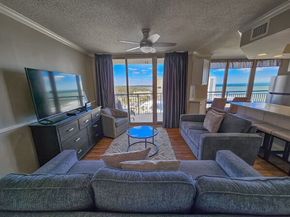 A modern living room with a flat-screen TV, grey sofas, a coffee table, and a balcony with a view of the city and ocean. - 1707 Brighton, impressively remodeled, 3 bedroom ocean view 3 Condo (Myrtle Beach)