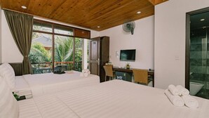 Habitación de hotel con dos camas, un escritorio, televisor y balcón con vista a palmeras.