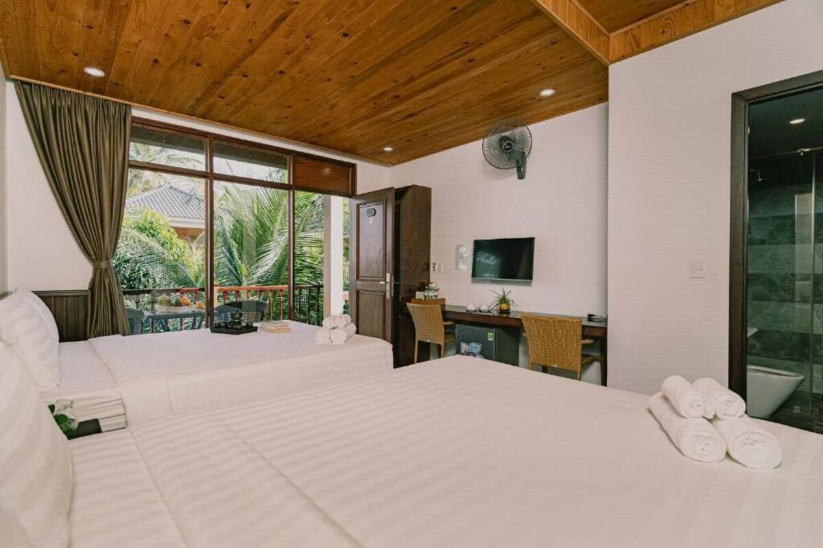 A hotel room with two beds, a desk, a television, and a balcony with a view of palm trees.