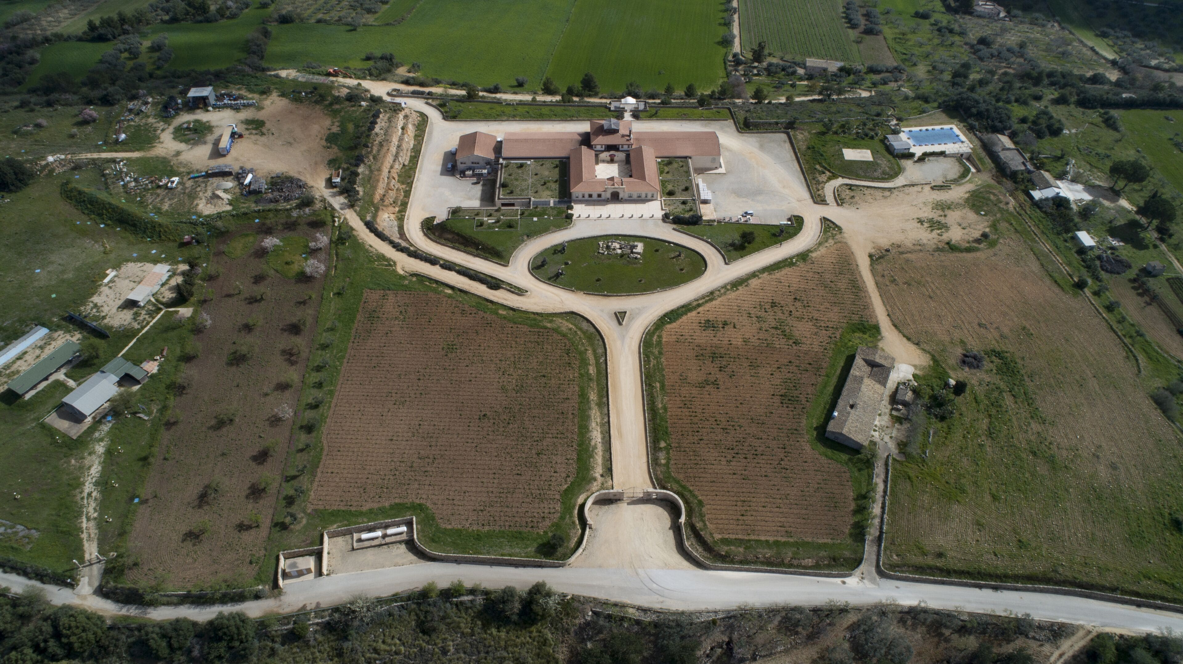 Una vista aerea di una grande tenuta con un edificio centrale circondato da campi aperti, un vialetto a senso unico e diverse strutture più piccole.