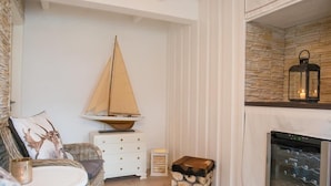 A cozy room with a wicker sofa, a small sailboat decoration, a stack of logs, and a wine refrigerator. - Strandhotel Lodge am Meer (Gluecksburg)