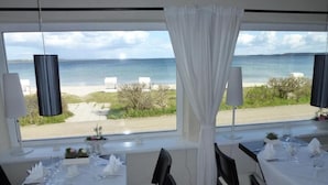 A dining area with tables set for two, large windows with a view of the sea and beach, and a white curtain partially drawn. - Strandhotel Lodge am Meer (Gluecksburg)
