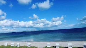 A beach with white lounge chairs aligned along the shoreline, a clear sky, and a calm sea. - Strandhotel Lodge am Meer (Gluecksburg)