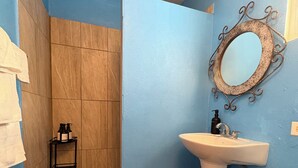 Un baño con pared azul, un lavamanos blanco, un espejo con marco decorativo, una zona de ducha y un estante pequeño con artículos de aseo.