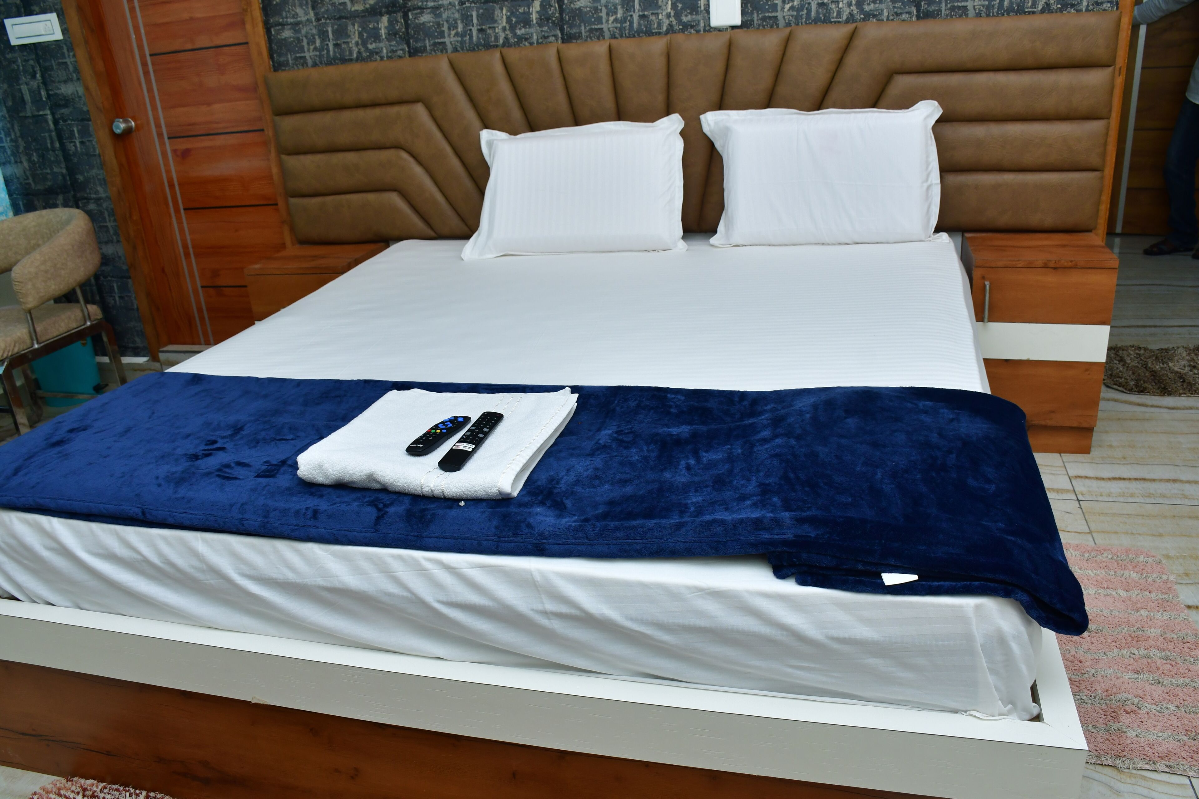 A neatly made bed with white and blue bedding, a brown headboard, and a remote control on the bed.