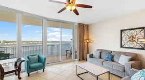 Amplia sala de estar con balcón, ventilador de techo y ventana grande con vistas al agua.