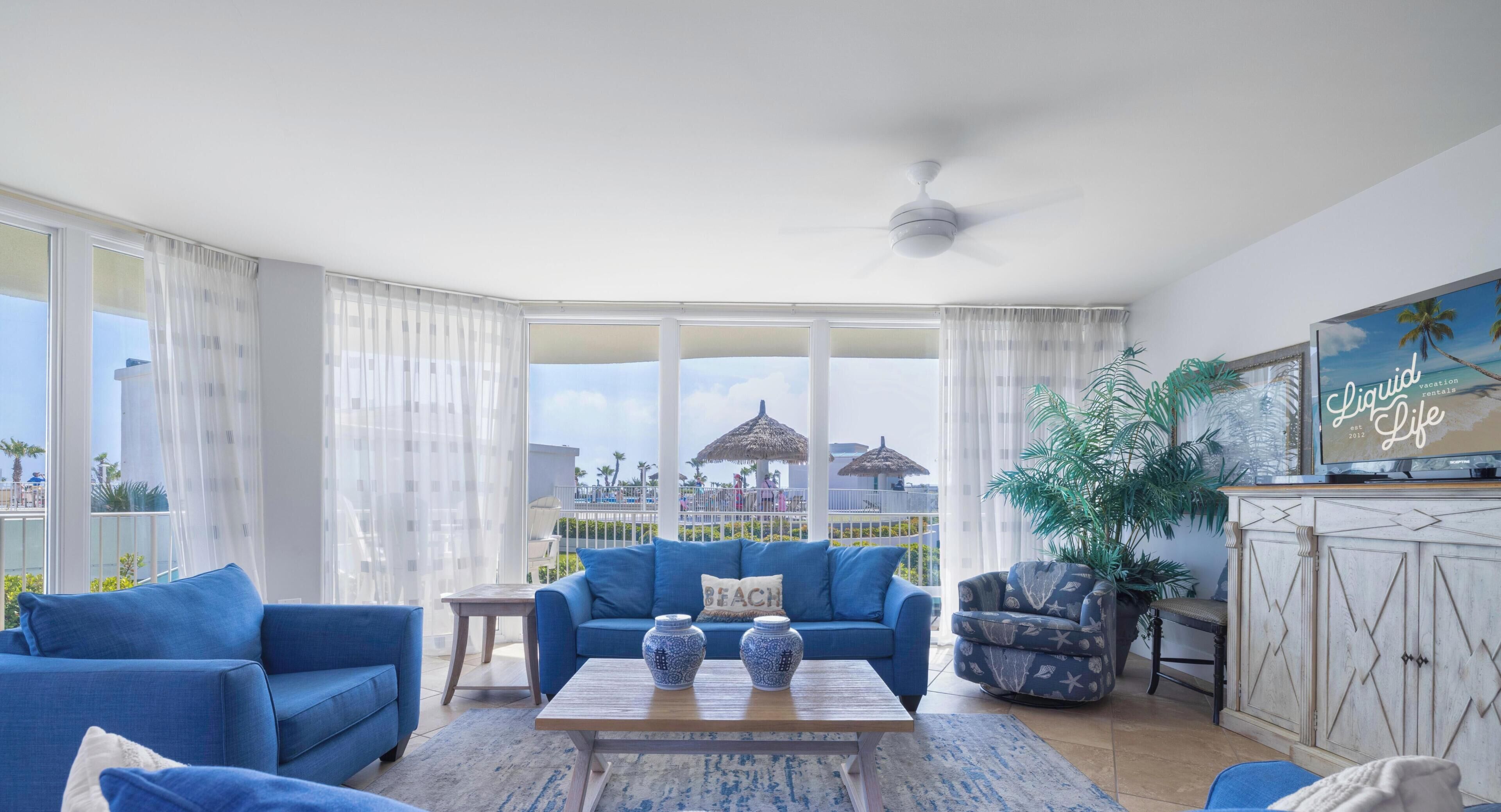 Een kamer met een blauwe bank, een bijzettafeltje met vazen, een plafondventilator en uitzicht op een strand met parasols.
