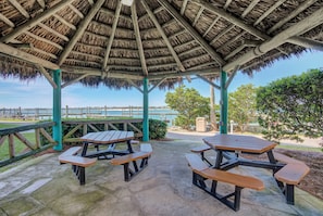 A covered picnic area with tables and benches, a thatched roof, and a view of the sea.