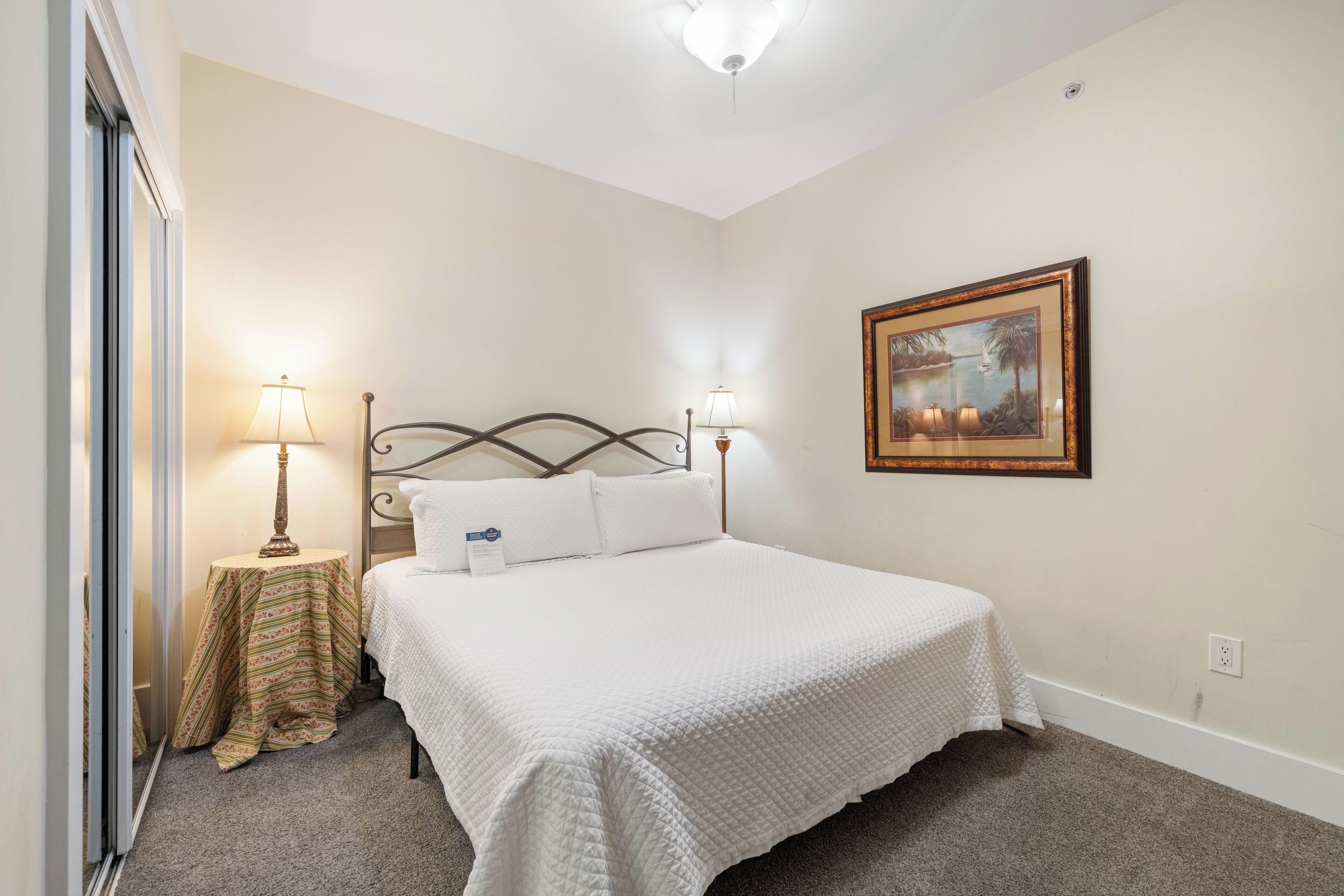 A neatly arranged bedroom with a bed, two bedside lamps, a small table with a lamp, and a framed picture on the wall.