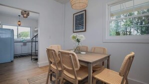 A dining area with a wooden table and wicker chairs, a hanging light fixture, and a window with a view of trees. - Ocean Boulevard North, 3 min walk to beach! (Rye)