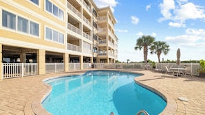 A swimming pool surrounded by a multi-story building with balconies, a covered patio area with seating, and palm trees in the background. - Marlin Key 3E by Liquid Life (Orange Beach)