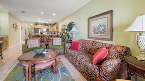A living room with a sofa, a coffee table, and a dining table. There is a kitchen in the background with cabinets and a refrigerator. - Marlin Key 3E by Liquid Life (Orange Beach)