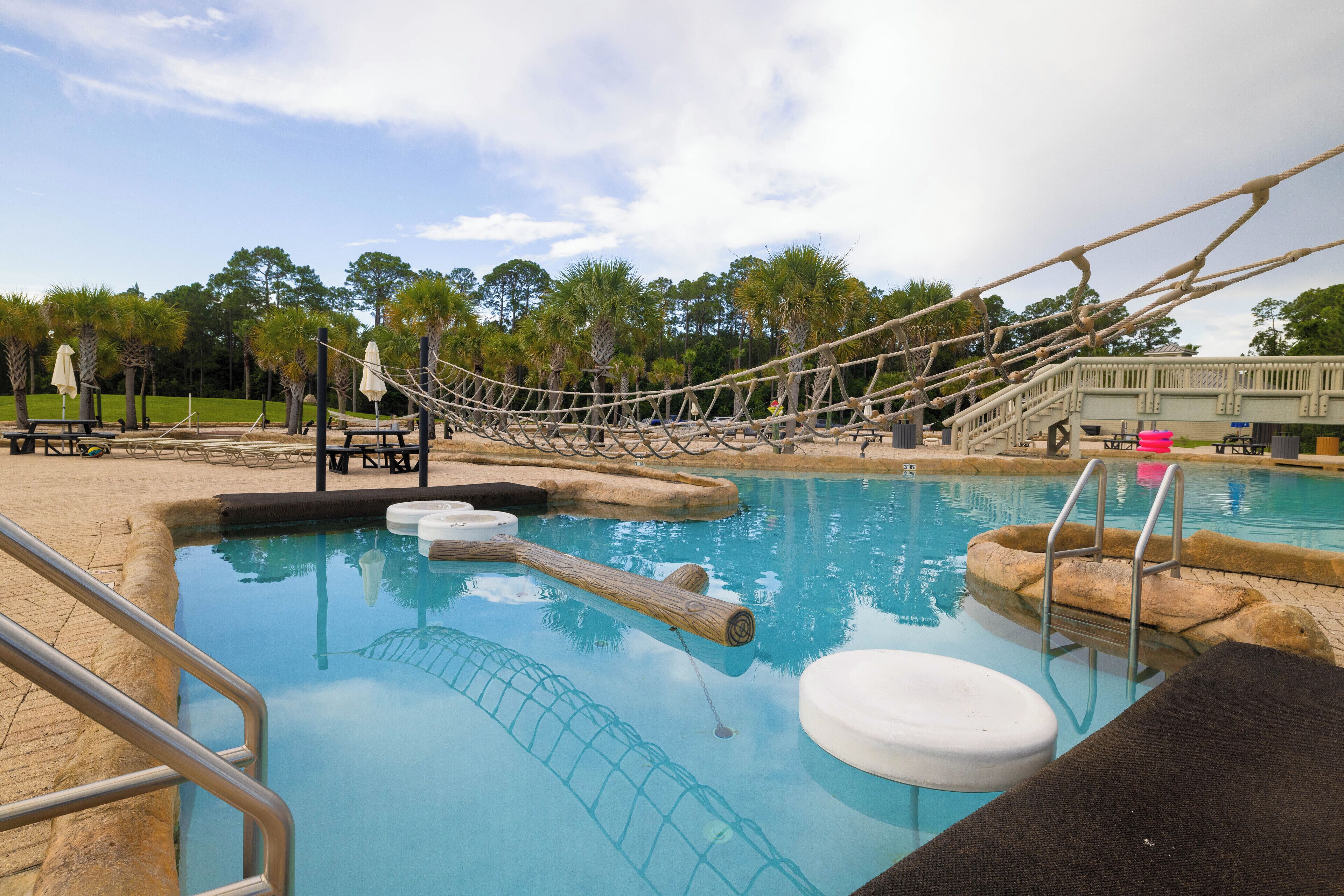 A pool with a rope bridge, lounge chairs, and a small waterfall feature.