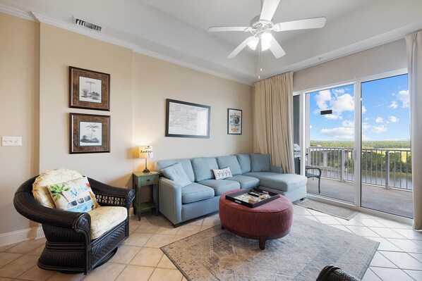 Un salon comprenant un canapé, un fauteuil, un pouf et un ventilateur de plafond. Des tableaux encadrés ornent le mur, et une porte coulissante offre une vue sur l’extérieur.