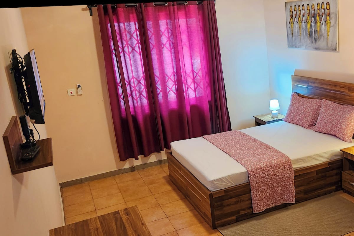 Un dormitorio con una cama de madera, una mesita de noche con una lámpara, un televisor sobre un mueble bajo y un cuadro en la pared.