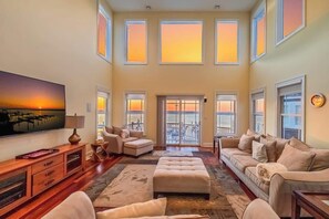 A spacious living room with a large flat-screen TV, multiple seating areas, and large windows with a view.