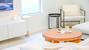 Un salon moderne avec un canapé blanc, une table basse ronde en bois ornée de bougies, un téléviseur fixé au mur et une fenêtre avec des stores.