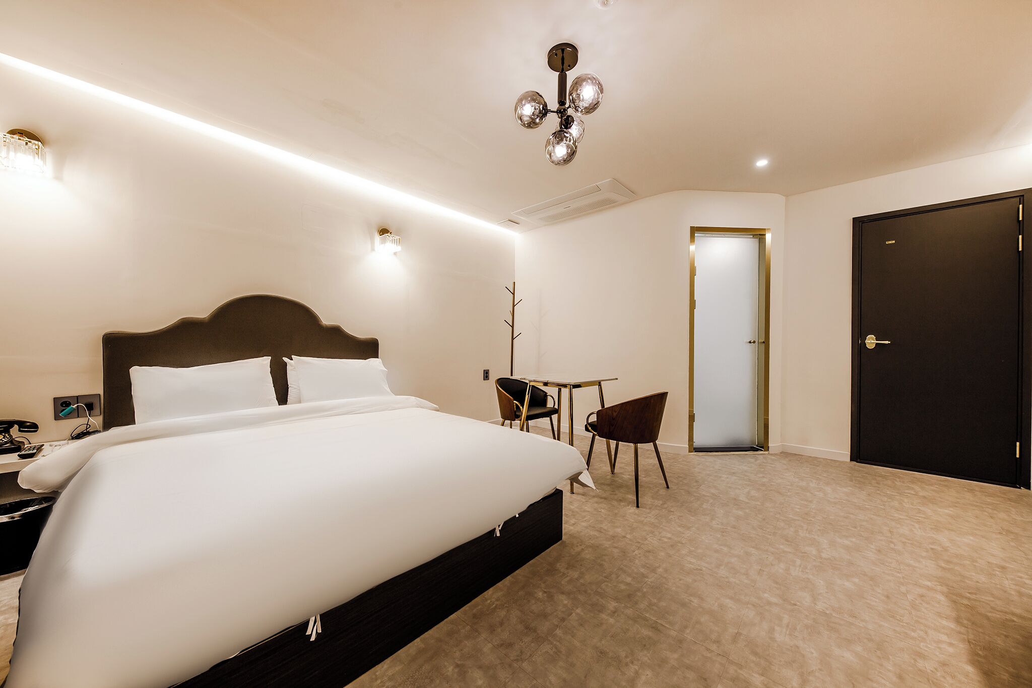 Quarto de hotel com uma cama grande, uma pequena mesa com duas cadeiras, uma porta escura e um lustre no teto.