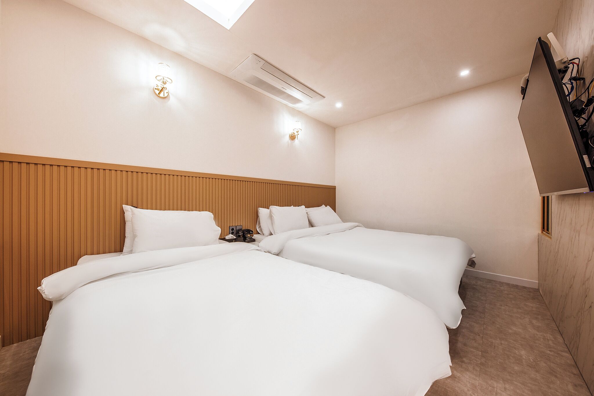 A hotel room with two beds, a wooden headboard, a ceiling-mounted air conditioner, and a wall-mounted TV.