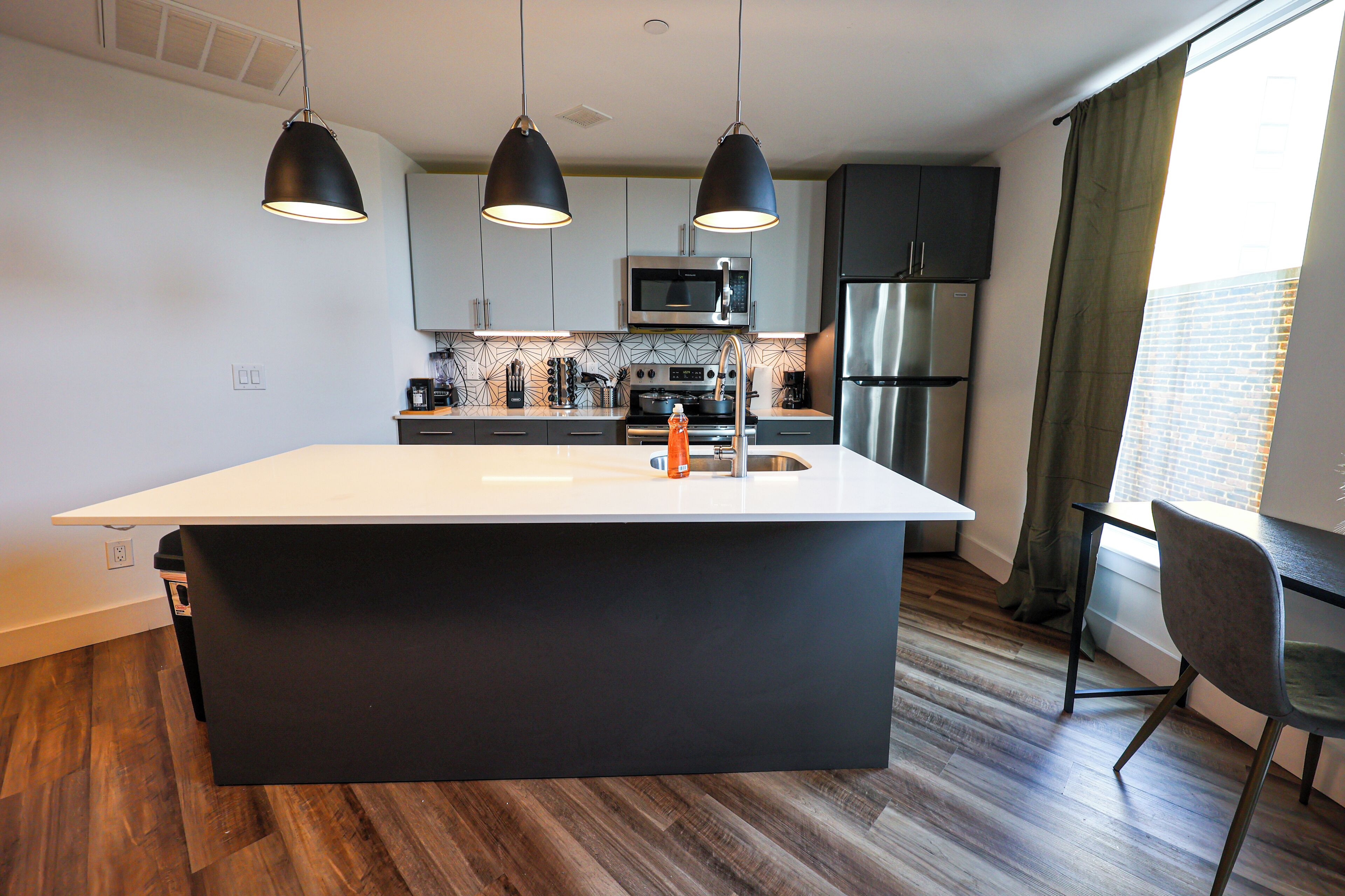 A modern kitchen with a central island, black pendant lights, stainless steel appliances, and a view of a brick building through a window.