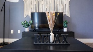 Una mesa negra con la palabra 'RELAX' en letras metálicas, un florero con plantas secas, un tapiz de macramé, un televisor y dos plantas en macetas.