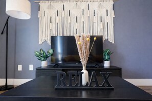 Un tavolo nero con la scritta 'RELAX' in metallo, un vaso con piante essiccate, un pannello decorativo in macramè, un televisore e due piante in vaso.