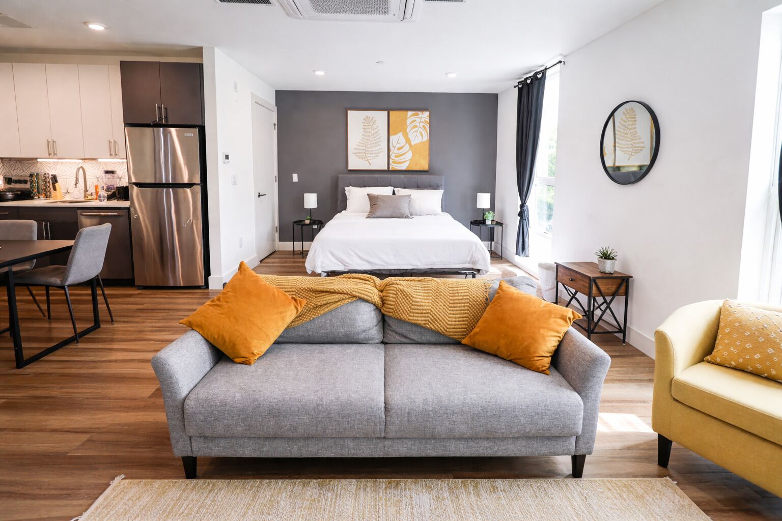 Une chambre d’hôtel moderne avec un canapé gris, un fauteuil jaune, un lit avec une literie blanche, un coin cuisine et un mur d’accent gris agrémenté d’œuvres d’art.