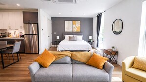 Una habitación de hotel moderna con un sofá gris, un sillón amarillo, una cama con ropa de cama blanca, una zona de cocina y una pared de acento gris con obras de arte.